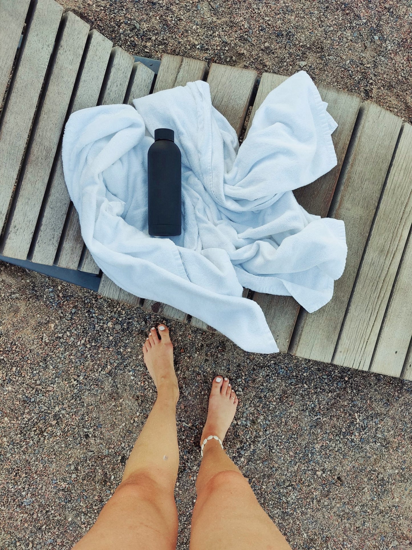 Musta terästä oleva juomapullo valkoisen pyyheen päällä. Alla pitkospuut ja hiekkaa. Edessä naisen jalat