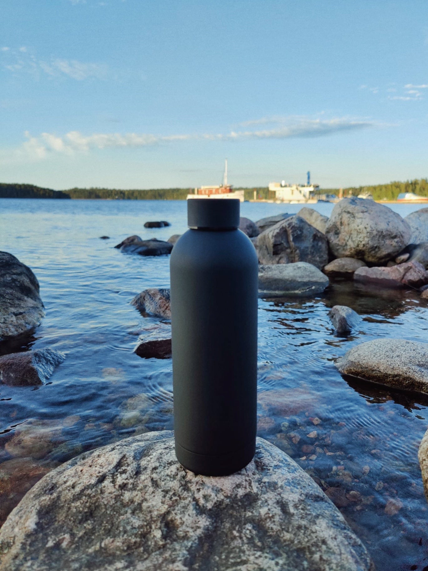 Musta teräsjuomapullo kiven päällä. Taustalla vesi maisema ja suuria kiviä