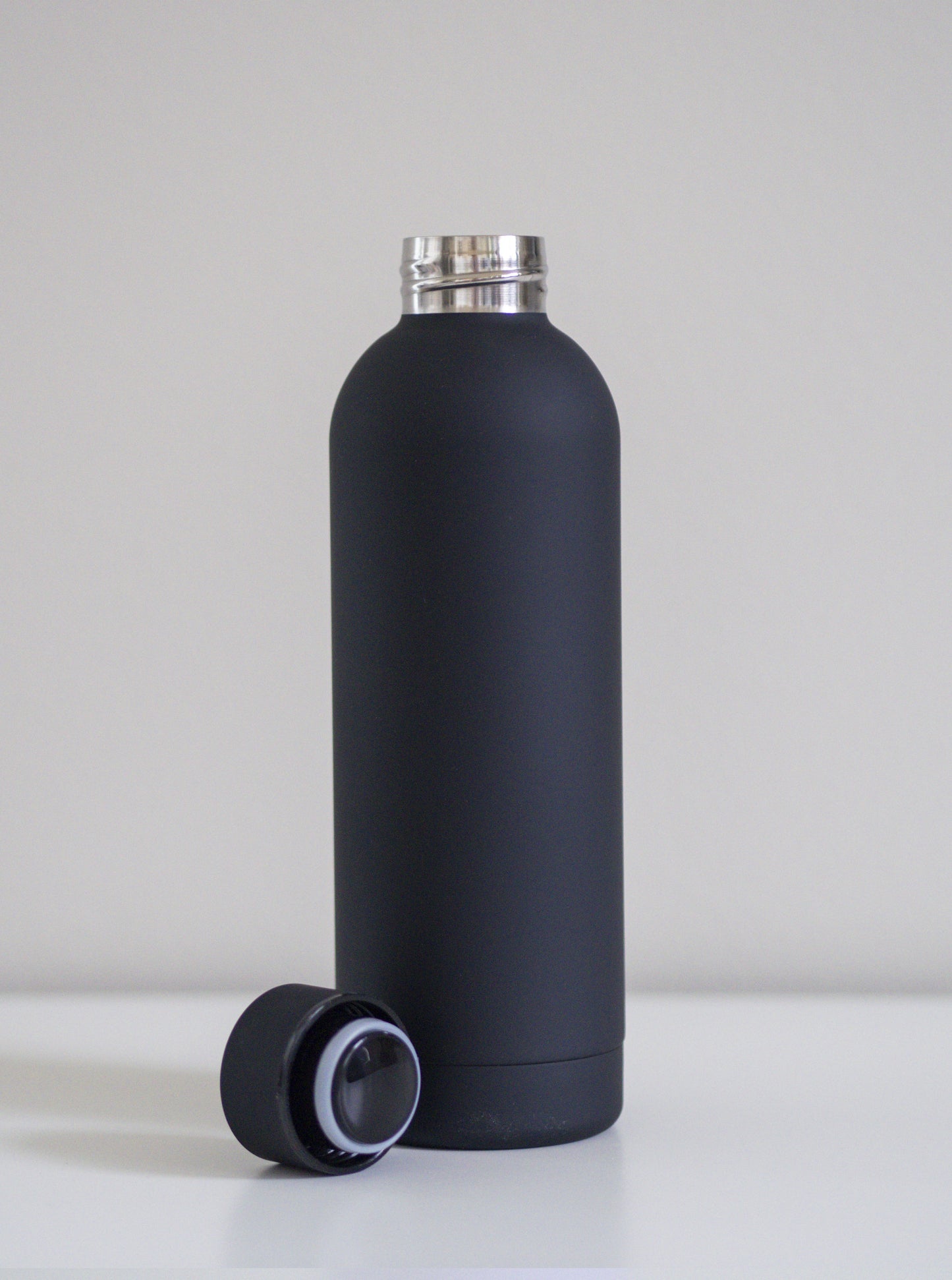 Musta ruostumatonta terästä oleva juomapullo ja korkki vieressä. Valkoista taustaa vasten